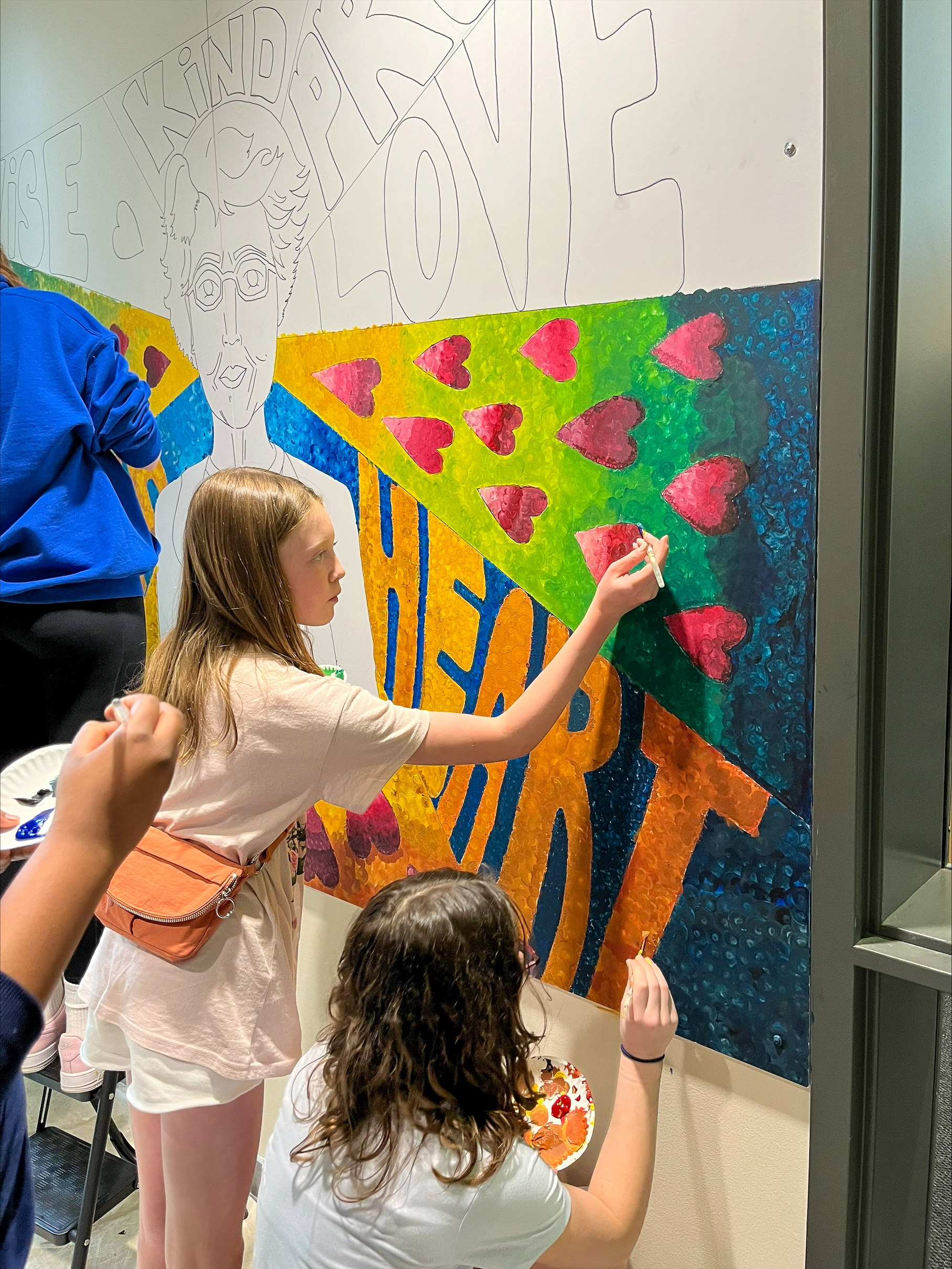 A young girl draws on a mural surrounded by her peers.